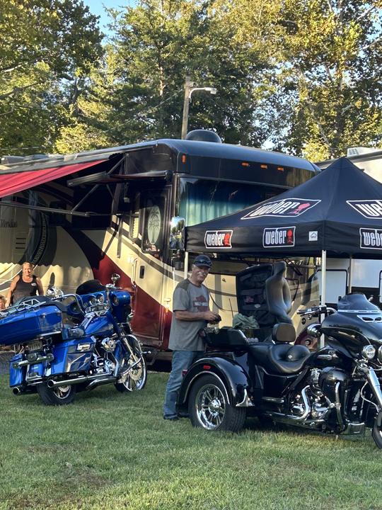 Touring motorcycles and a trike parked on the grass beside a motorhome, a rider standing with them