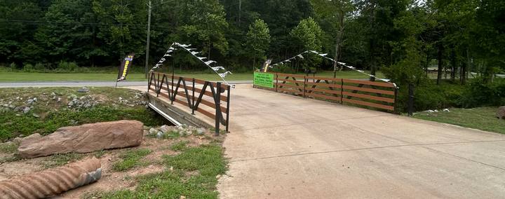 Wide view of a paved drive crossing a bridge with wooden rails, checkered flags strung overhead, rocks and logs along the bank