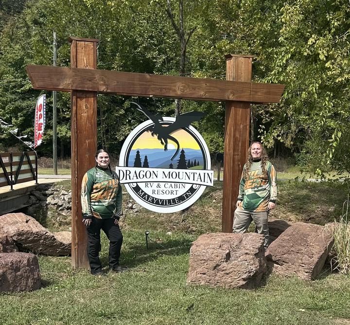 Two people standing beside the Dragon Mountain RV & Cabin Resort entrance sign, which hangs from a timber frame among boulders