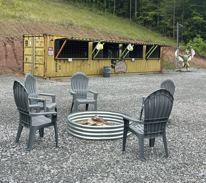 Adirondack chairs around a fire ring on gravel, a converted shipping container with a service window behind