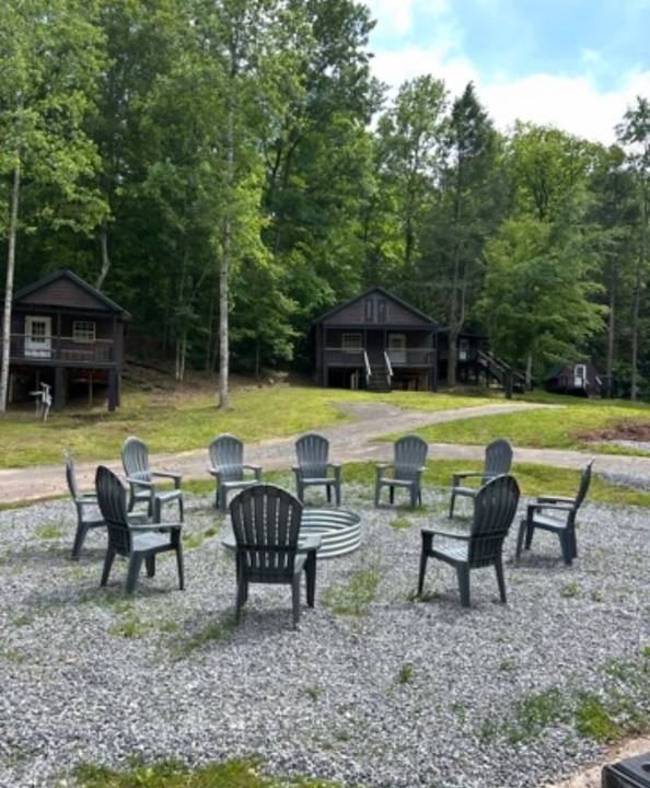 A circle of Adirondack chairs around a fire ring in a clearing, cabins in the trees behind