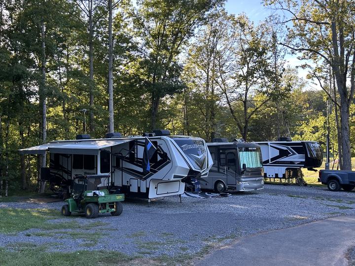 Two fifth wheels and a motorhome parked in a row on gravel, a green utility cart out front