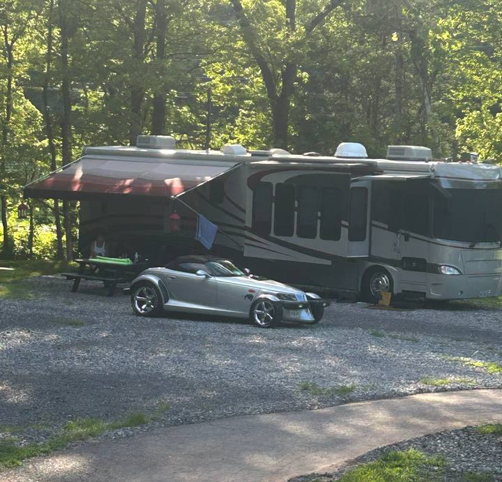 A large motorhome and a sports car parked on a gravel site in the trees
